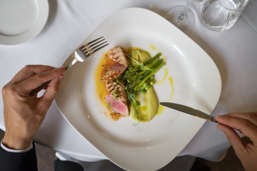Comensal corta un filete de pescado servido con espárragos y puré en un plato blanco, vista cenital sobre mantel blanco